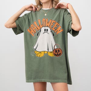 May include: Olive green Halloween t-shirt featuring a cartoon ghost character with a sad expression, holding a jack-o'-lantern bucket. The word "HALLOWEEN" is arched above the ghost in orange letters. The ghost has yellow feet.