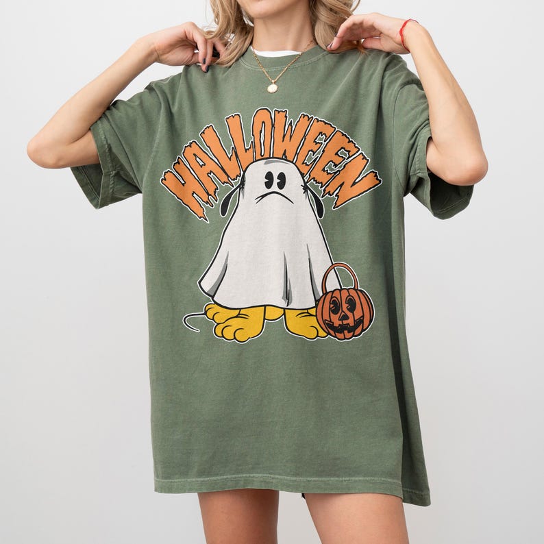 May include: Olive green Halloween t-shirt featuring a cartoon ghost character with a sad expression, holding a jack-o'-lantern bucket. The word "HALLOWEEN" is arched above the ghost in orange letters. The ghost has yellow feet.