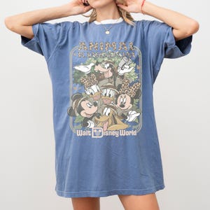 May include: Blue t-shirt featuring a graphic of Disney characters in safari attire, with the text "ANIMAL KINGDOM" and "Walt Disney World". The design includes Mickey Mouse, Minnie Mouse, Donald Duck, Goofy, and Pluto.