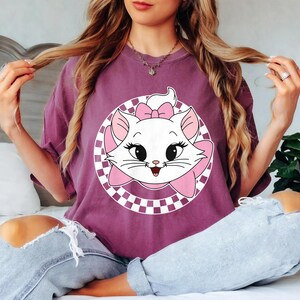 May include: A mauve t-shirt with a white cat graphic, pink bow, and checkered circle design. The cat has black eyes and a smiling face. The shirt is worn by a person.
