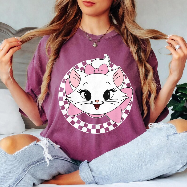 May include: A mauve t-shirt with a white cat graphic, pink bow, and checkered circle design. The cat has black eyes and a smiling face. The shirt is worn by a person.