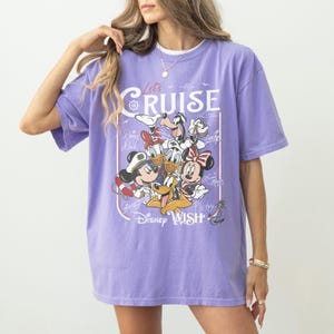 May include: A lilac-colored t-shirt featuring a graphic of Disney characters, including Mickey Mouse, Minnie Mouse, Donald Duck, and Goofy. The shirt has the words "Let's Cruise" and "Disney Wish" printed on it.