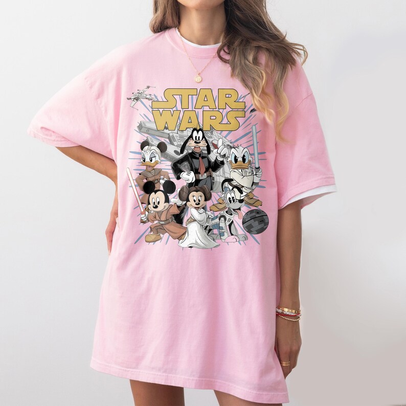 May include: Pink t-shirt featuring a graphic of Star Wars characters, including Mickey Mouse, Minnie Mouse, Donald Duck, and Goofy. The words "STAR WARS" are printed in yellow across the top of the design.