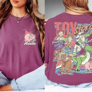 K&ouml;nnte beinhalten: Pflaumenfarbenes T-Shirt mit Toy Story-Motiv. Auf der Vorderseite befindet sich eine Schweinegrafik und das Wort "Auntie". Auf der R&uuml;ckseite sind das Toy Story-Logo und Charaktere wie Woody und Buzz Lightyear abgebildet.