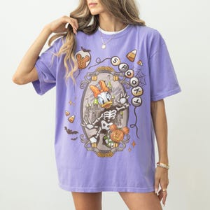 May include: A lilac-colored t-shirt featuring a Halloween-themed graphic. The design includes Daisy Duck in a skeleton costume, a castle, and the words "Spookt" and "Daisy Duck". Additional elements include bats, candy corn, and a Mickey Mouse pumpkin.
