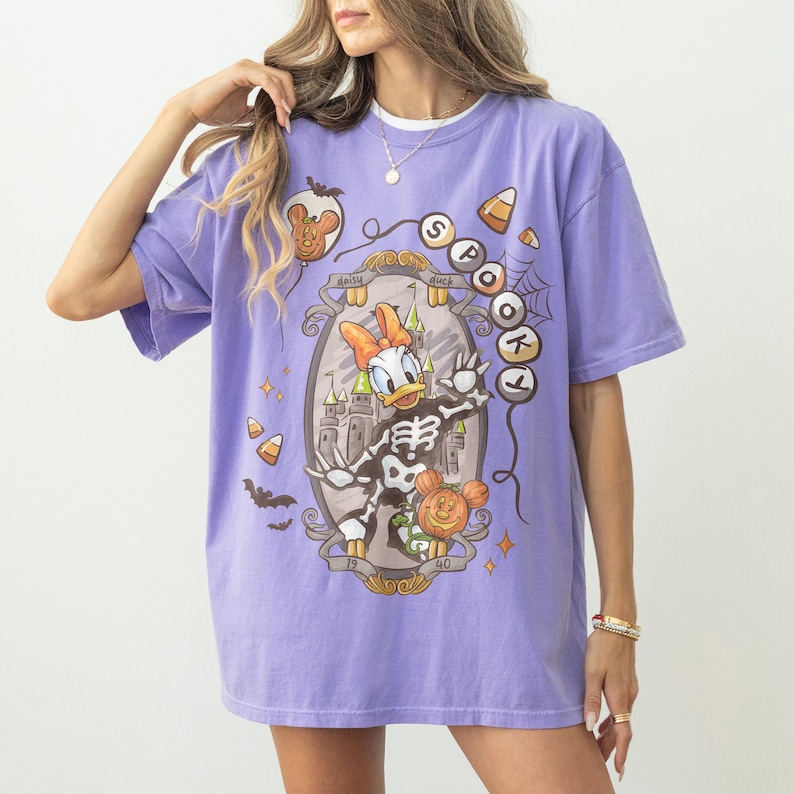 May include: A lilac-colored t-shirt featuring a Halloween-themed graphic. The design includes Daisy Duck in a skeleton costume, a castle, and the words "Spookt" and "Daisy Duck". Additional elements include bats, candy corn, and a Mickey Mouse pumpkin.