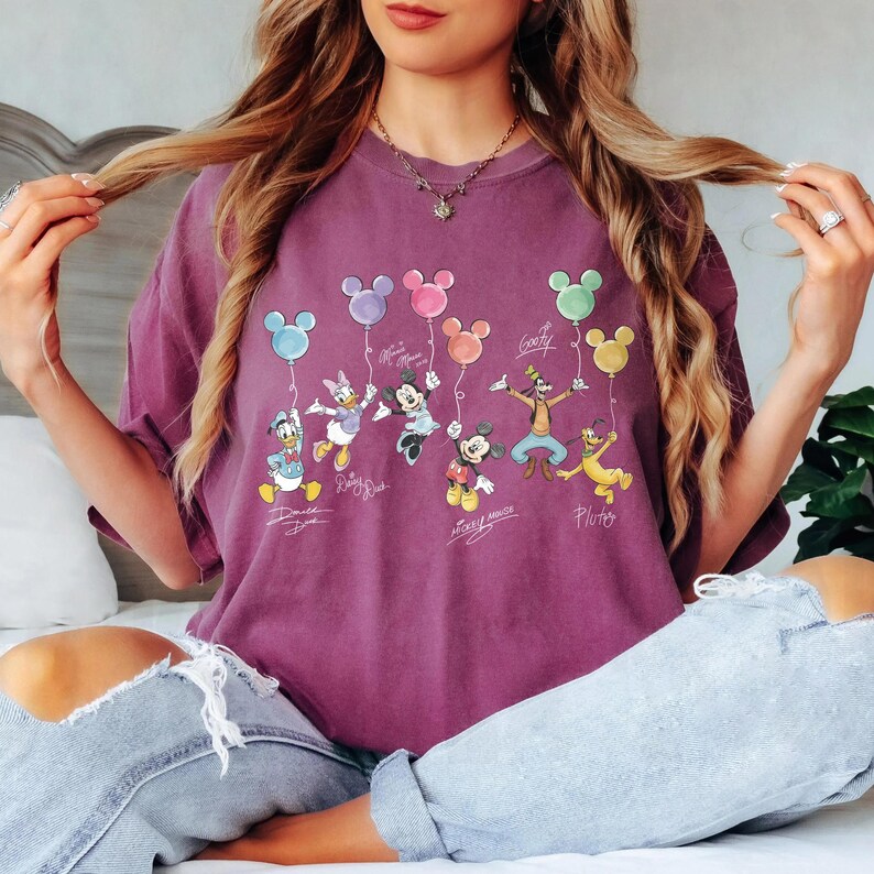 May include: A purple t-shirt featuring Disney characters holding colorful Mickey Mouse balloons. The characters include Donald Duck, Daisy Duck, Minnie Mouse, Mickey Mouse, Goofy, and Pluto. The shirt has a vintage look.