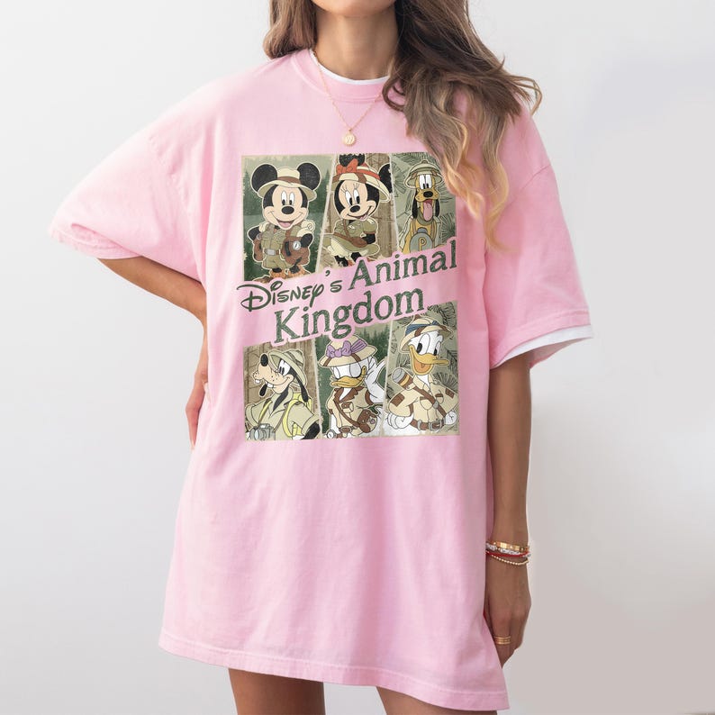 May include: A light pink t-shirt featuring a graphic of Disney's Animal Kingdom characters, including Mickey Mouse, Minnie Mouse, Goofy, Donald Duck, and Pluto. The shirt has a white trim on the sleeves and collar.