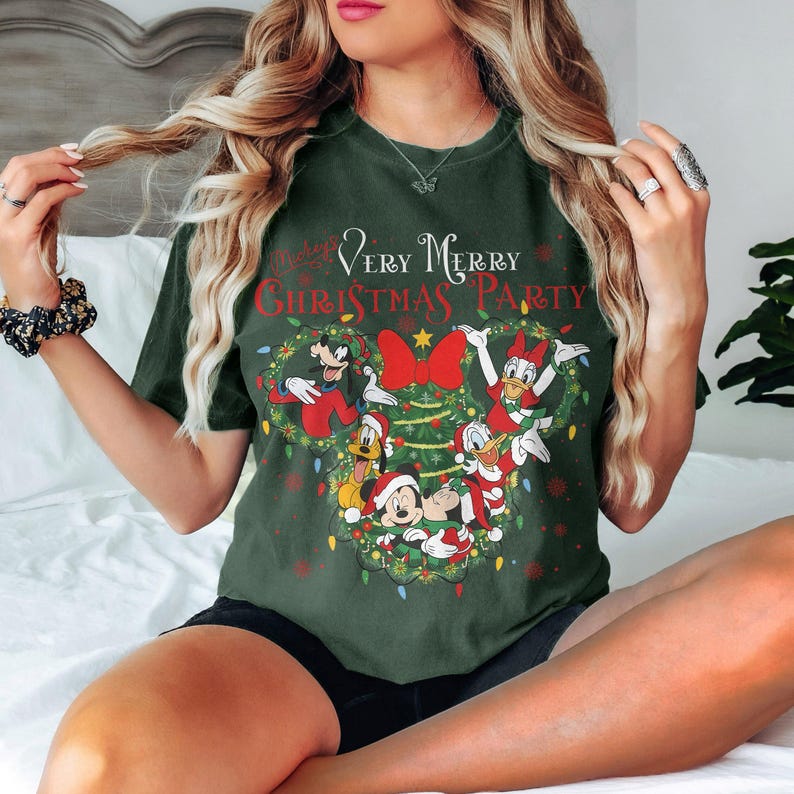 May include: Dark green t-shirt featuring a festive design with Mickey Mouse, Minnie Mouse, Donald Duck, Goofy, and Pluto. The text "Very Merry Christmas Party" is displayed above the characters, surrounded by a wreath of lights and a red bow.