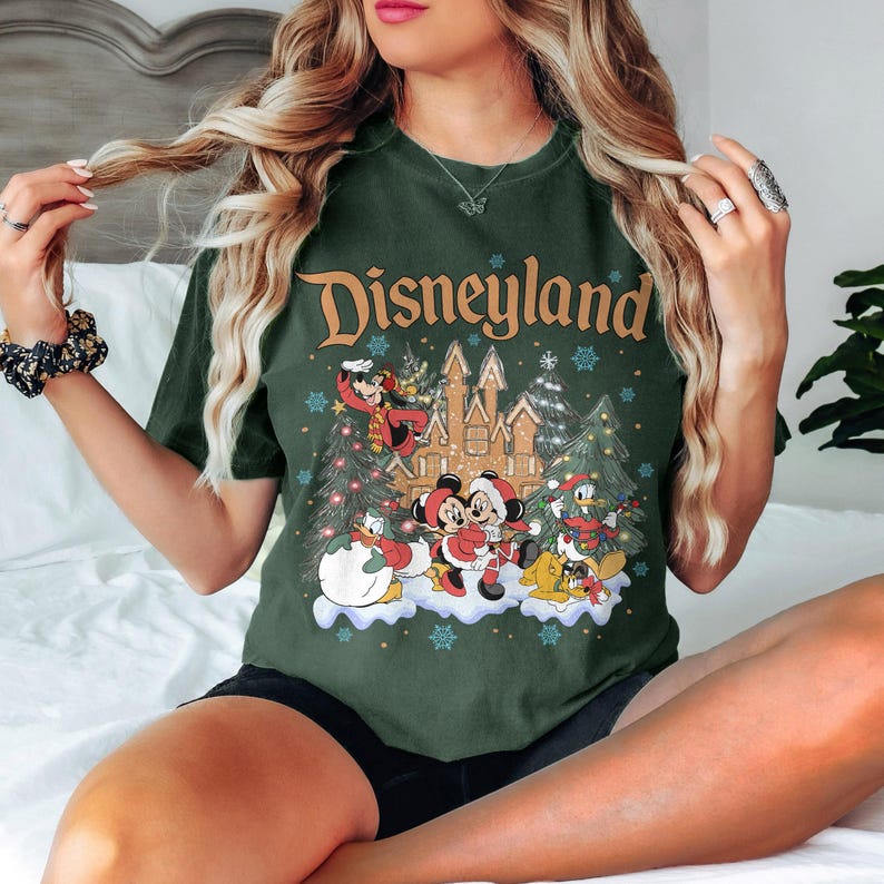 K&ouml;nnte beinhalten: Dunkelgr&uuml;nes T-Shirt mit dem Wort "Disneyland" in goldener Schrift. Das Shirt zeigt eine festliche Grafik von Disney-Figuren, darunter Mickey und Minnie Mouse, in einer Winterszene mit einem Schloss und Weihnachtsb&auml;umen.
