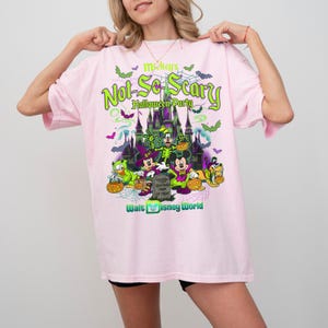 May include: A light pink t-shirt featuring a graphic of Mickey's Not-So-Scary Halloween Party at Walt Disney World. The design includes a castle, bats, spiderwebs, and characters in costumes holding pumpkins, with text in green and purple.