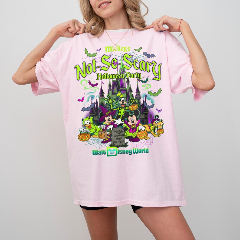 May include: A light pink t-shirt featuring a graphic of Mickey's Not-So-Scary Halloween Party at Walt Disney World. The design includes a castle, bats, spiderwebs, and characters in costumes holding pumpkins, with text in green and purple.