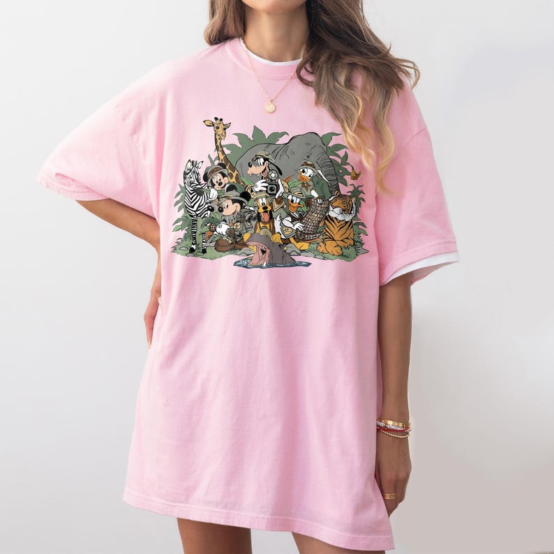 K&ouml;nnte beinhalten: Hellrosa T-Shirt mit einem farbenfrohen Aufdruck von Zeichentrickfiguren und Tieren, darunter ein Zebra, eine Giraffe, ein Elefant, ein Tiger und Mickey Mouse. Das Design ist vor einem Hintergrund aus gr&uuml;nem Laub angeordnet.