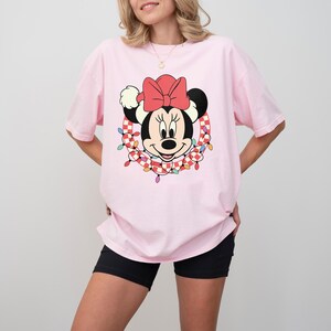 May include: A light pink t-shirt featuring a Minnie Mouse graphic. The graphic shows Minnie's face with a red bow, surrounded by a wreath of checkered garland and colorful lights. The shirt has short sleeves and a relaxed fit.