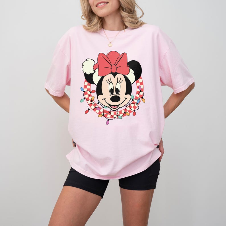 May include: A light pink t-shirt featuring a Minnie Mouse graphic. The graphic shows Minnie's face with a red bow, surrounded by a wreath of checkered garland and colorful lights. The shirt has short sleeves and a relaxed fit.