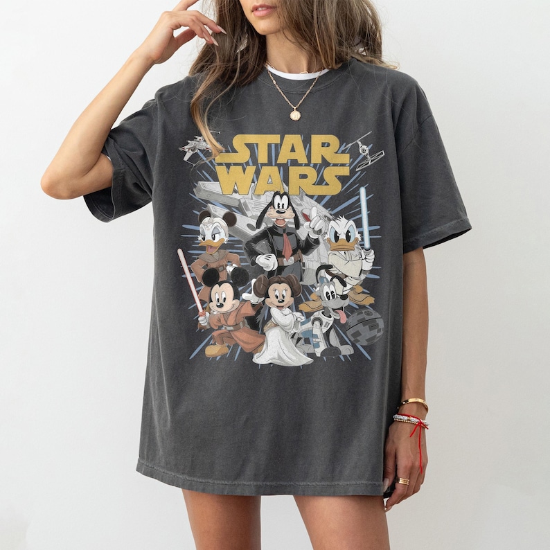 May include: Dark gray t-shirt featuring a Star Wars graphic with cartoon characters. The design includes the Star Wars logo in gold, and various Disney characters dressed as Star Wars characters, such as Mickey Mouse and Donald Duck.