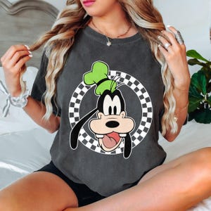 May include: Dark grey t-shirt with a cartoon image of Goofy wearing a green hat, set against a black and white chequered circle. The graphic is in black, white, and green. The t-shirt is a casual style.