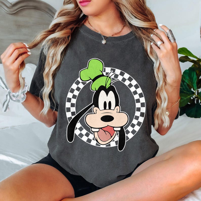 May include: Dark grey t-shirt with a cartoon image of Goofy wearing a green hat, set against a black and white chequered circle. The graphic is in black, white, and green. The t-shirt is a casual style.