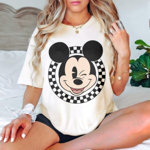 May include: A cream-coloured t-shirt with a large graphic of Mickey Mouse winking. The design includes a black and white chequered circle around Mickey's face. The shirt is a casual, short-sleeved style.
