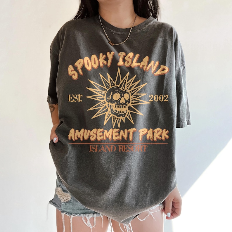 May include: Dark gray t-shirt with the text "SPOOKY ISLAND AMUSEMENT PARK ISLAND RESORT EST. 2002" in orange and yellow. The graphic features a skull with a sunburst design. The shirt is oversized.