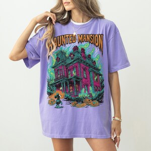 May include: A lilac-colored t-shirt featuring a colorful graphic of a haunted mansion with the text "HAUNTED MANSION" in orange. The graphic includes ghosts, pumpkins, and a cartoon character. The shirt has short sleeves and a relaxed fit.