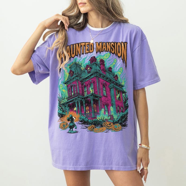 May include: A lilac-colored t-shirt featuring a colorful graphic of a haunted mansion with the text "HAUNTED MANSION" in orange. The graphic includes ghosts, pumpkins, and a cartoon character. The shirt has short sleeves and a relaxed fit.