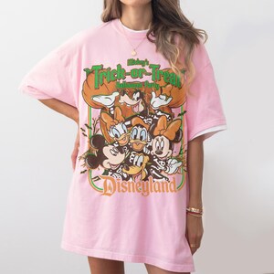 K&ouml;nnte beinhalten: Ein hellrosa T-Shirt mit kurzen &Auml;rmeln und einem Grafikdruck von Mickey Maus, Minnie Maus, Donald Duck, Daisy Duck und Goofy. Der Text lautet Mickey's Trick-or-Treat Halloween Party und Disneyland, umgeben von K&uuml;rbissen und Flederm&auml;usen.