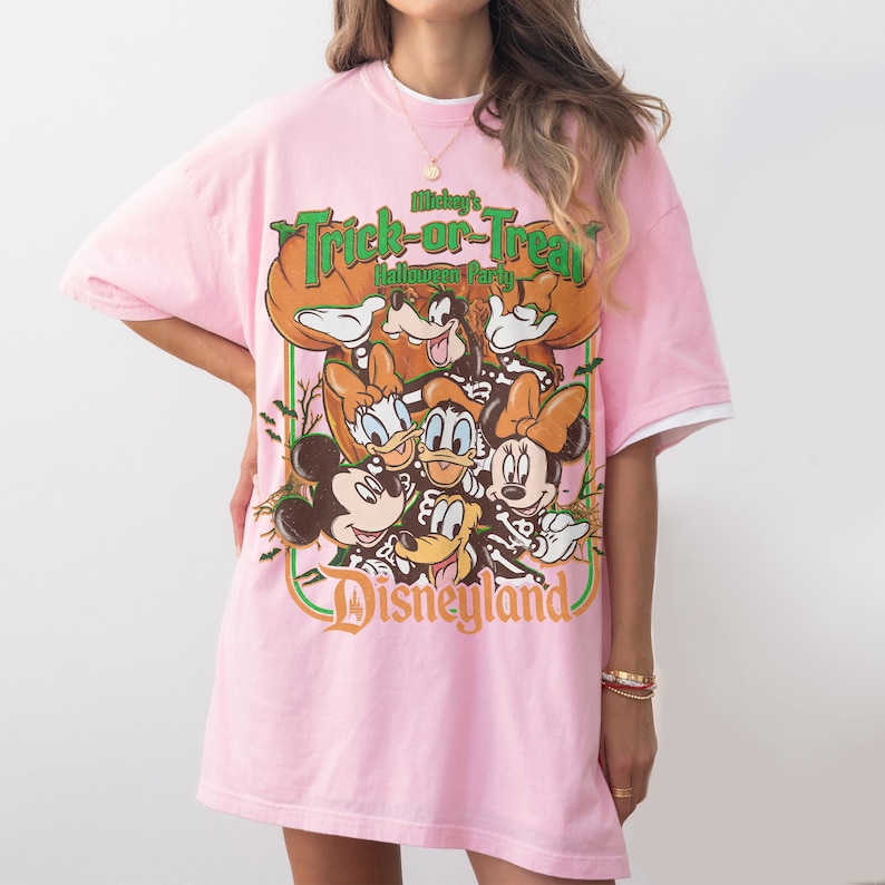 K&ouml;nnte beinhalten: Ein hellrosa T-Shirt mit kurzen &Auml;rmeln und einem Grafikdruck von Mickey Maus, Minnie Maus, Donald Duck, Daisy Duck und Goofy. Der Text lautet Mickey's Trick-or-Treat Halloween Party und Disneyland, umgeben von K&uuml;rbissen und Flederm&auml;usen.
