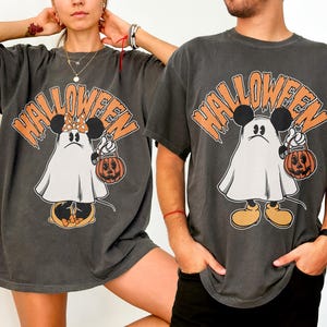 May include: Dark gray Halloween t-shirts featuring a cartoon ghost character with Mickey Mouse ears holding a jack-o'-lantern. The word "HALLOWEEN" is printed above the character in orange. The shirts have short sleeves and a relaxed fit.