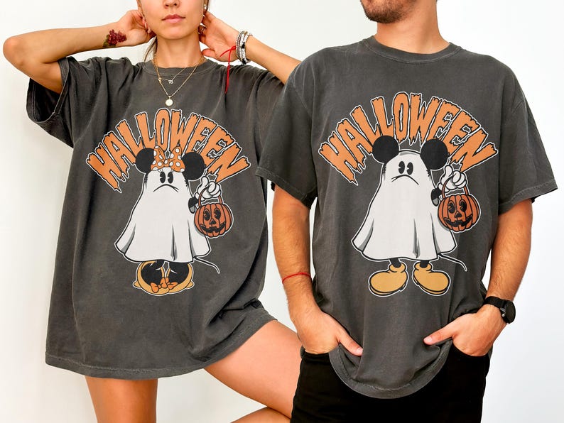 May include: Dark gray Halloween t-shirts featuring a cartoon ghost character with Mickey Mouse ears holding a jack-o'-lantern. The word "HALLOWEEN" is printed above the character in orange. The shirts have short sleeves and a relaxed fit.