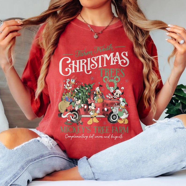 May include: Red t-shirt with a graphic featuring Mickey Mouse and friends on a red truck carrying a Christmas tree. The text reads "Farm Fresh Christmas Trees" and "Mickey's Tree Farm".