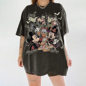 Może przedstawiać: Ciemnoszary t-shirt o kroju oversize z grafiką o tematyce Halloween, przedstawiającą postacie Disneya, takie jak Myszka Miki, Myszka Minnie, Goofy, Kaczor Donald, Kaczka Daisy i Pluto. Nadruk zawiera pajęczyny, nietoperze oraz napisy kursywą.