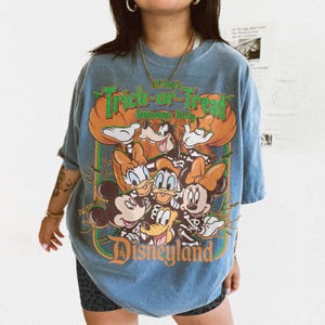 K&ouml;nnte beinhalten: Ein blaues Kurzarm-T-Shirt mit einem Grafikdruck von Disney-Figuren in Skelettkost&uuml;men. Der Text lautet Mickey's Trick-or-Treat Halloween Party und Disneyland. Das Design enth&auml;lt K&uuml;rbisse, Flederm&auml;use sowie eine lebendige orangefarbene und gr&uuml;ne Farbpalette.