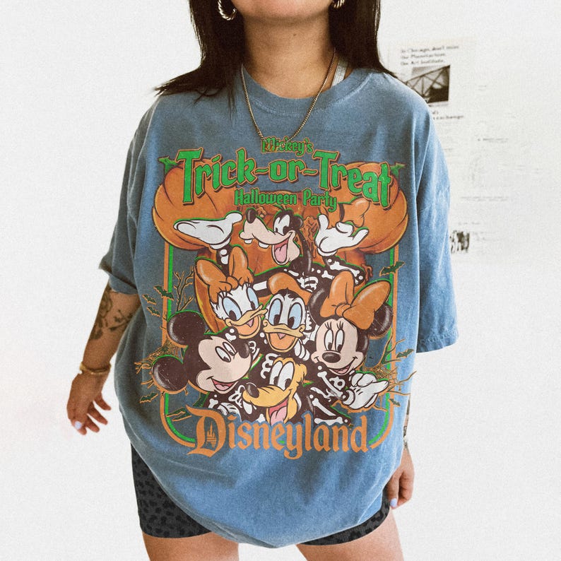 K&ouml;nnte beinhalten: Ein blaues Kurzarm-T-Shirt mit einem Grafikdruck von Disney-Figuren in Skelettkost&uuml;men. Der Text lautet Mickey's Trick-or-Treat Halloween Party und Disneyland. Das Design enth&auml;lt K&uuml;rbisse, Flederm&auml;use sowie eine lebendige orangefarbene und gr&uuml;ne Farbpalette.