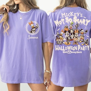 May include: A light purple t-shirt featuring a Halloween-themed design. The front displays a cartoon of Daisy Duck in a skeleton costume, with the name "Joanna". The back has a larger graphic with Mickey Mouse and friends in skeleton costumes, with the text "Mickey's Not So Scary Halloween Party".