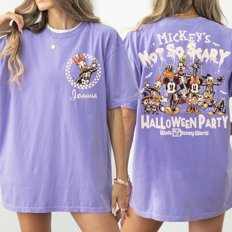 May include: A light purple t-shirt featuring a Halloween-themed design. The front displays a cartoon of Daisy Duck in a skeleton costume, with the name "Joanna". The back has a larger graphic with Mickey Mouse and friends in skeleton costumes, with the text "Mickey's Not So Scary Halloween Party".