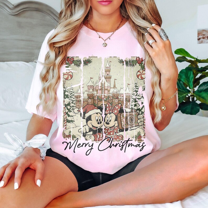 May include: A light pink t-shirt featuring a distressed graphic of Mickey and Minnie Mouse in front of a castle, with the words "Merry Christmas" in black script. The design includes Christmas trees, candy canes, and snow. The shirt has short sleeves.