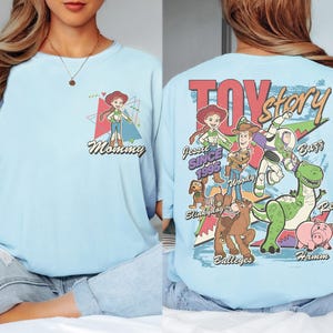 K&ouml;nnte beinhalten: Hellblaues T-Shirt mit einem Aufdruck von Jessie aus Toy Story und dem Wort "Mommy" auf der Vorderseite. Auf der R&uuml;ckseite des Shirts sind das Toy Story-Logo und Charaktere wie Woody, Buzz und Rex abgebildet. Der Text "Since 1995" ist ebenfalls aufgedruckt.