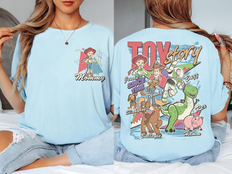 K&ouml;nnte beinhalten: Hellblaues T-Shirt mit einem Aufdruck von Jessie aus Toy Story und dem Wort "Mommy" auf der Vorderseite. Auf der R&uuml;ckseite des Shirts sind das Toy Story-Logo und Charaktere wie Woody, Buzz und Rex abgebildet. Der Text "Since 1995" ist ebenfalls aufgedruckt.
