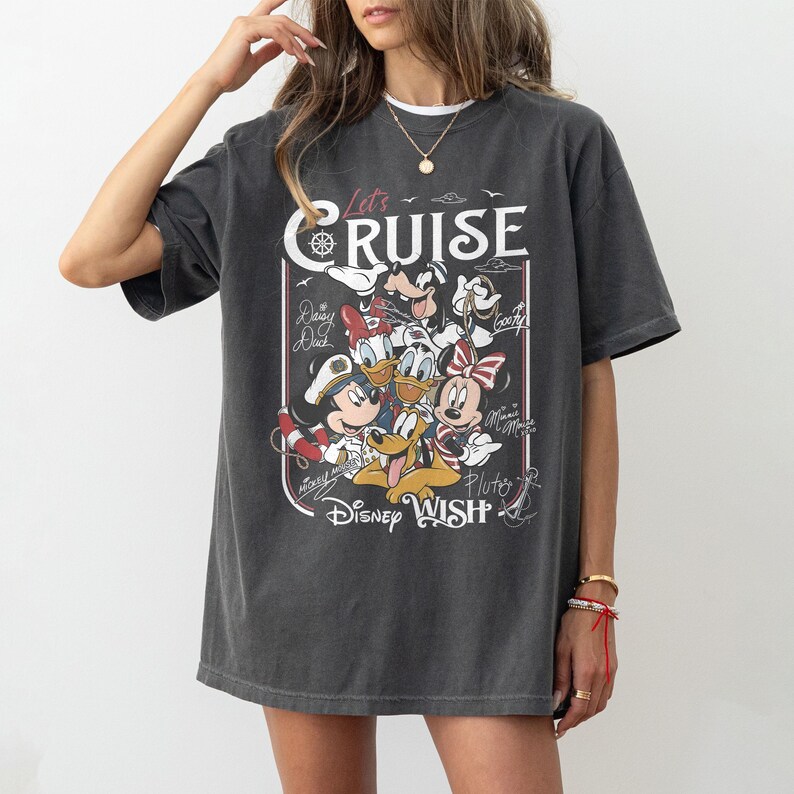May include: Dark gray t-shirt featuring a graphic of Disney characters, including Mickey Mouse, Minnie Mouse, Donald Duck, and Pluto. The shirt has the text "Let's Cruise" and "Disney Wish" in a vintage style.