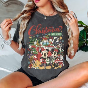 May include: Dark gray t-shirt featuring a festive Christmas design with Mickey and Minnie Mouse, Donald Duck, and other Disney characters. The text "Christmas on Main Street" is displayed in red and green.