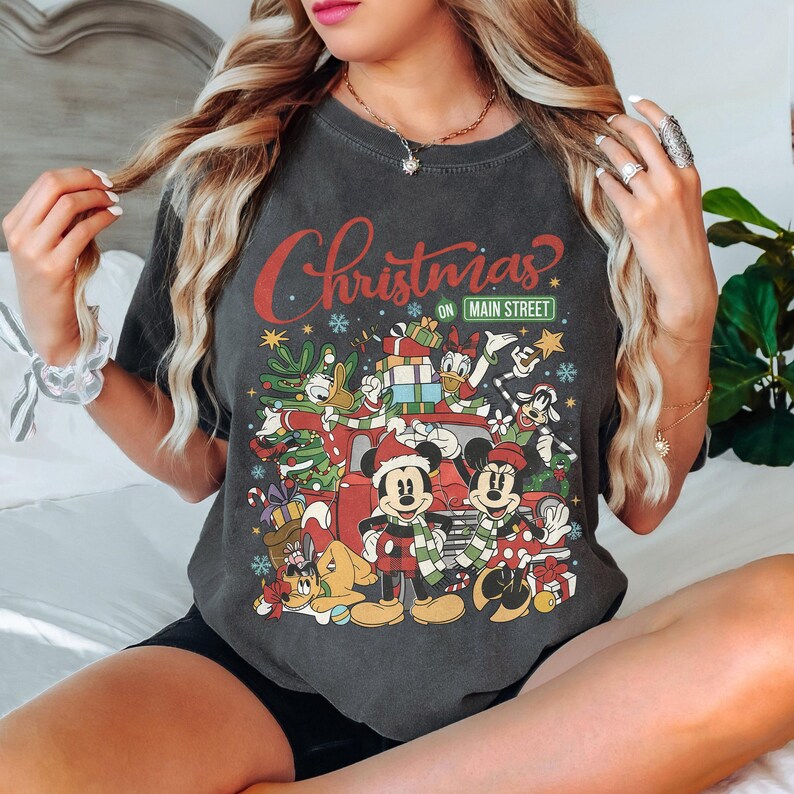 May include: Dark gray t-shirt featuring a festive Christmas design with Mickey and Minnie Mouse, Donald Duck, and other Disney characters. The text "Christmas on Main Street" is displayed in red and green.