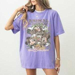 May include: A lilac-colored t-shirt featuring a graphic of Disney characters, including Mickey and Minnie Mouse, Donald and Daisy Duck, and Goofy. The text "ANIMAL KINGDOM" and "Walt Disney World" are also displayed on the shirt.