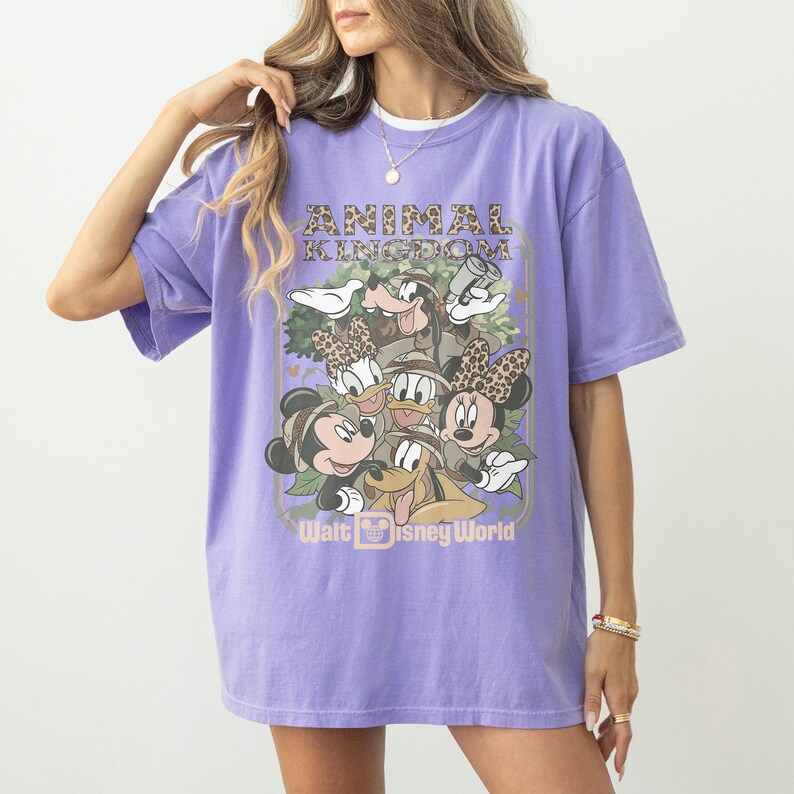 May include: A lilac-colored t-shirt featuring a graphic of Disney characters, including Mickey and Minnie Mouse, Donald and Daisy Duck, and Goofy. The text "ANIMAL KINGDOM" and "Walt Disney World" are also displayed on the shirt.