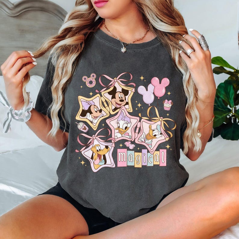 May include: Dark gray t-shirt featuring Disney characters Mickey, Minnie, Daisy, and Pluto in star-shaped frames. The word "magical" is displayed in pastel colors below the characters. Pink and yellow bows and balloons add to the whimsical design.