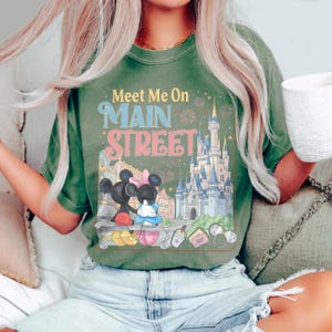 Op de afbeelding: Groen t-shirt met de tekst "Meet Me On Main Street" en een afbeelding van Mickey en Minnie Mouse voor een kasteel. Het shirt heeft een vintage, vervaagde uitstraling.