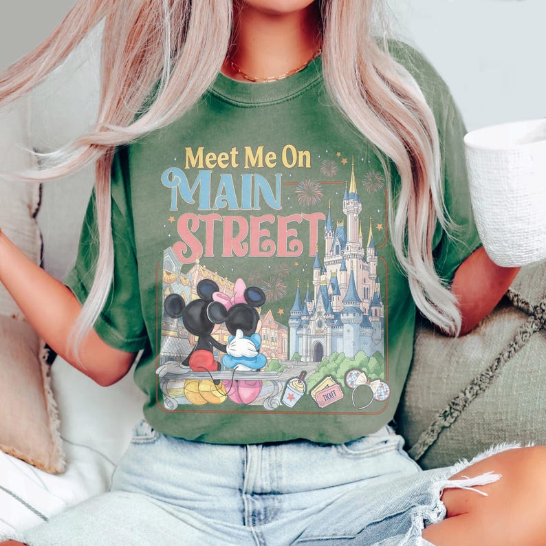 Op de afbeelding: Groen t-shirt met de tekst "Meet Me On Main Street" en een afbeelding van Mickey en Minnie Mouse voor een kasteel. Het shirt heeft een vintage, vervaagde uitstraling.