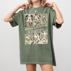 May include: Olive green t-shirt featuring a graphic of Disney's Animal Kingdom characters, including Mickey and Minnie Mouse, Goofy, Donald Duck, and Pluto. The design is in a vintage style, with the words "Disney's Animal Kingdom" printed on the front.
