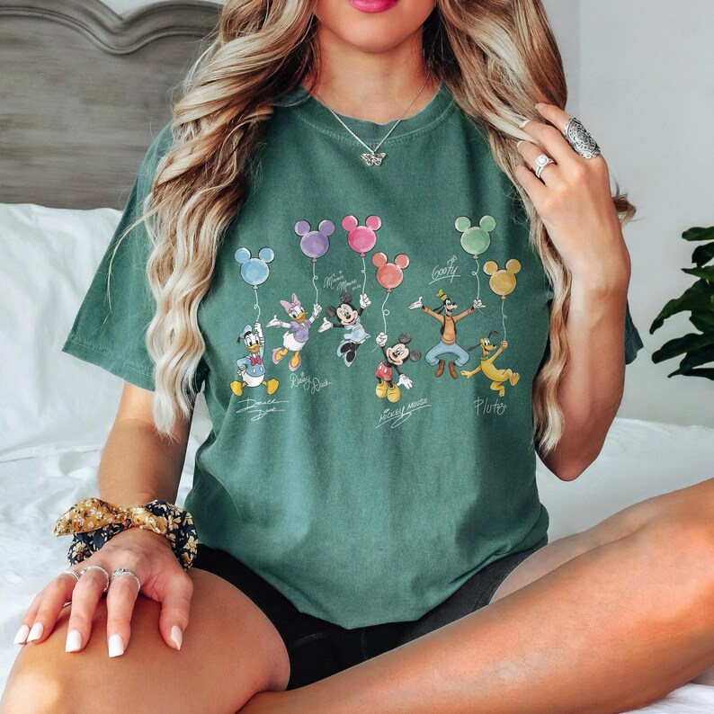 May include: Green t-shirt featuring Disney characters holding Mickey Mouse-shaped balloons. Characters include Donald Duck, Daisy Duck, Mickey Mouse, Minnie Mouse, Goofy, and Pluto. The shirt has the characters' signatures below them.
