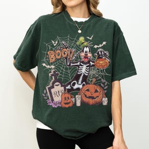 May include: A dark green short-sleeve t-shirt featuring a graphic of a cartoon dog in a skeleton costume, a tombstone with the text RIP, a jack-o'-lantern, and the word BOO. The design includes a spiderweb background with bats and candles.